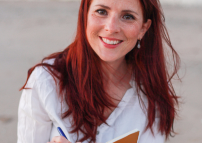 Charlotte TREMEL assise sur la plage, écrivant sur un carnet, symbolisant un moment de réflexion et de création pour les entrepreneurs et professionnels artistiques. BLOG